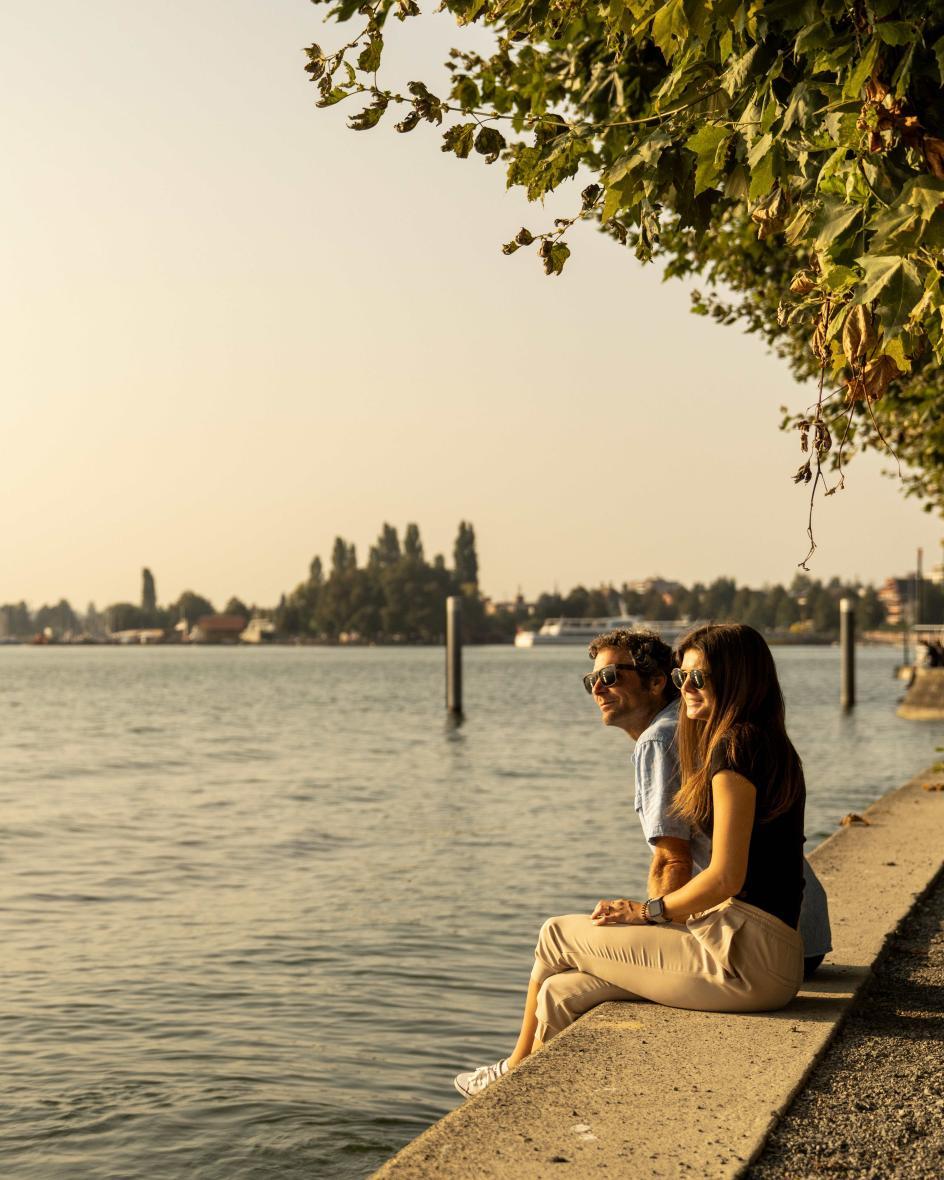 Zwei Menschen sitzen am Ufer des Zugersee und blicken in den goldgelben Sonnenuntergang.